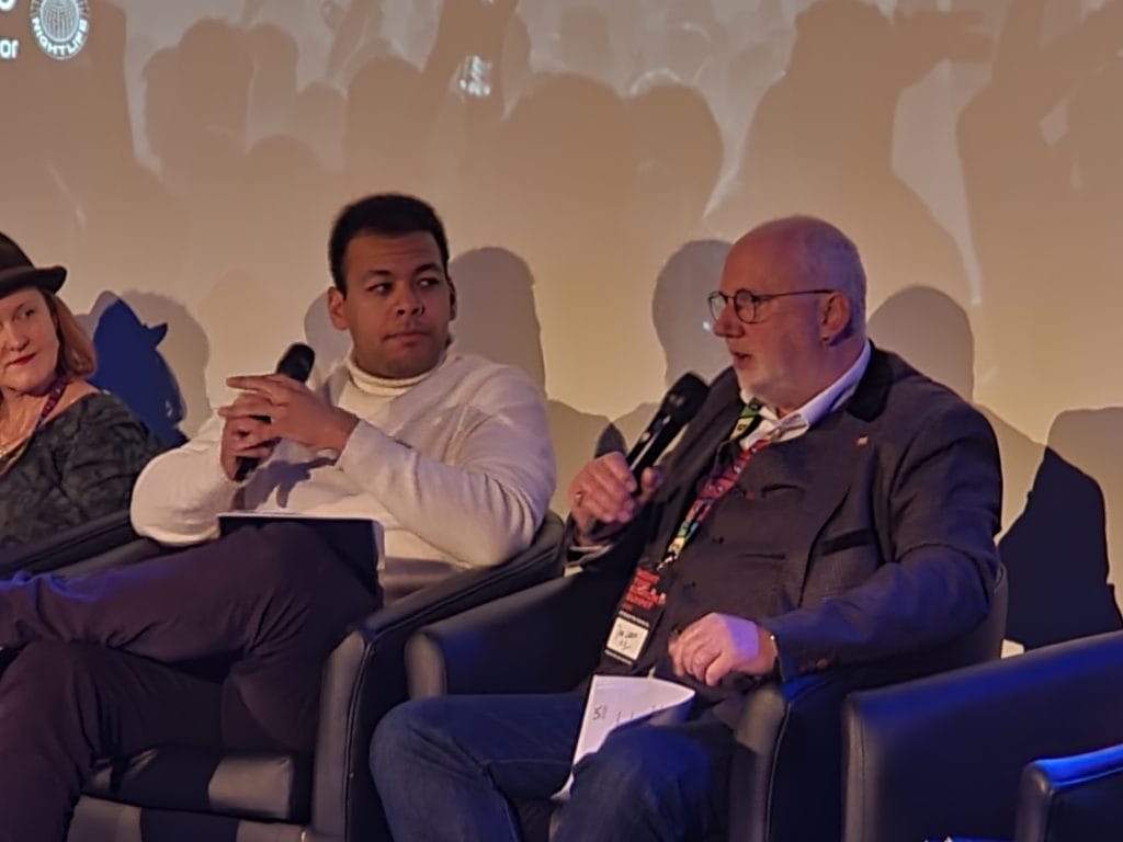 This is a photograph of Dave Webster talking into a microphone with Jamie Njoku-Goodwin listening intently.
