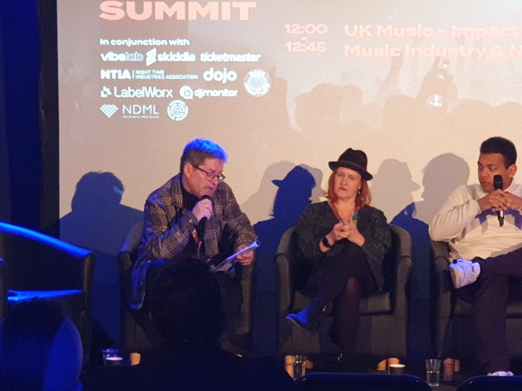 This is a picture of Kevin Brennan talking into a microphone seated in front of a large screen with Annabella Coldrick and Jamie Njoku-Goodwin to his side. 